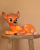 The Resin Deer Lamp features a cartoon-style fawn with big eyes lying on its side atop a wooden stool, creating a delightful decor accent for children's rooms, shown against a plain light brown background.