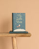 A copy of "The Little Prince | Collector's Edition | Hardcover" stands upright on a wooden bench, with another copy lying beneath it against a solid light beige background.