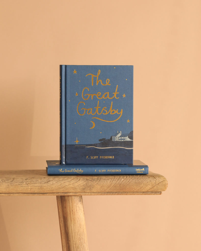 A blue hardcover Collector's Edition of "The Great Gatsby" stands upright on a wooden bench with another copy beneath it, set against a beige background. The cover displays gold stars and a seaside scene, highlighting this iconic novel.