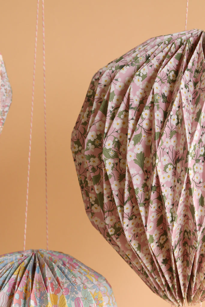 Close-up of Meri Meri x Liberty Origami Balloon Decorations featuring paper lanterns with Liberty pastel floral prints, hanging by pink and white strings against a peach background—ideal for baby showers.