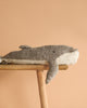 The Senger Naturwelt Cuddly Animal - Whale, a handmade plush toy of organic cotton with a gray top and white underside, sits on a wooden stool against a plain beige background.