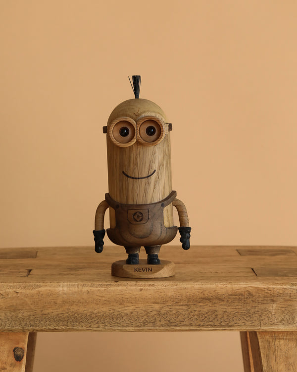 The Boyhood Minions Kevin, Small is a collectible wooden figurine crafted from European FSC White Oak, featuring Sir Kevin's goggles, black gloves, tuft of hair, and a smiling face, displayed on a wooden bench against a beige background.