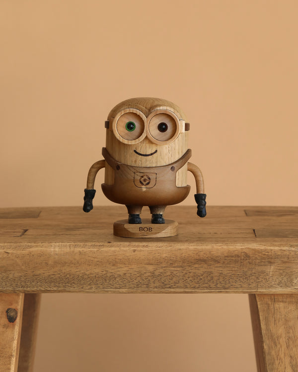 The Boyhood Minion Bob wooden figurine, featuring mismatched eyes and a "BOB" label on its base, stands on a wooden table with a plain beige background.