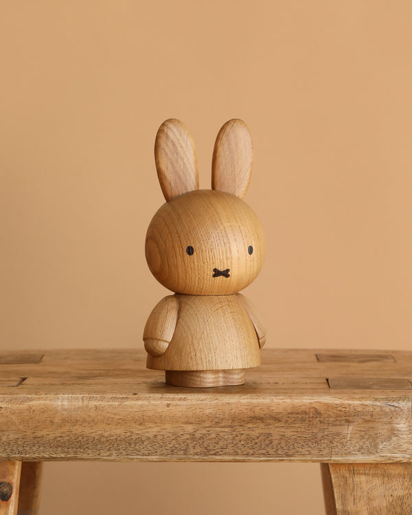 The Boyhood Miffy x Miffy, Small wooden collectible with simple black facial features sits on a wooden bench against a beige background.