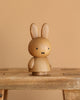 The Boyhood Miffy x Miffy, Small wooden collectible with simple black facial features sits on a wooden bench against a beige background.