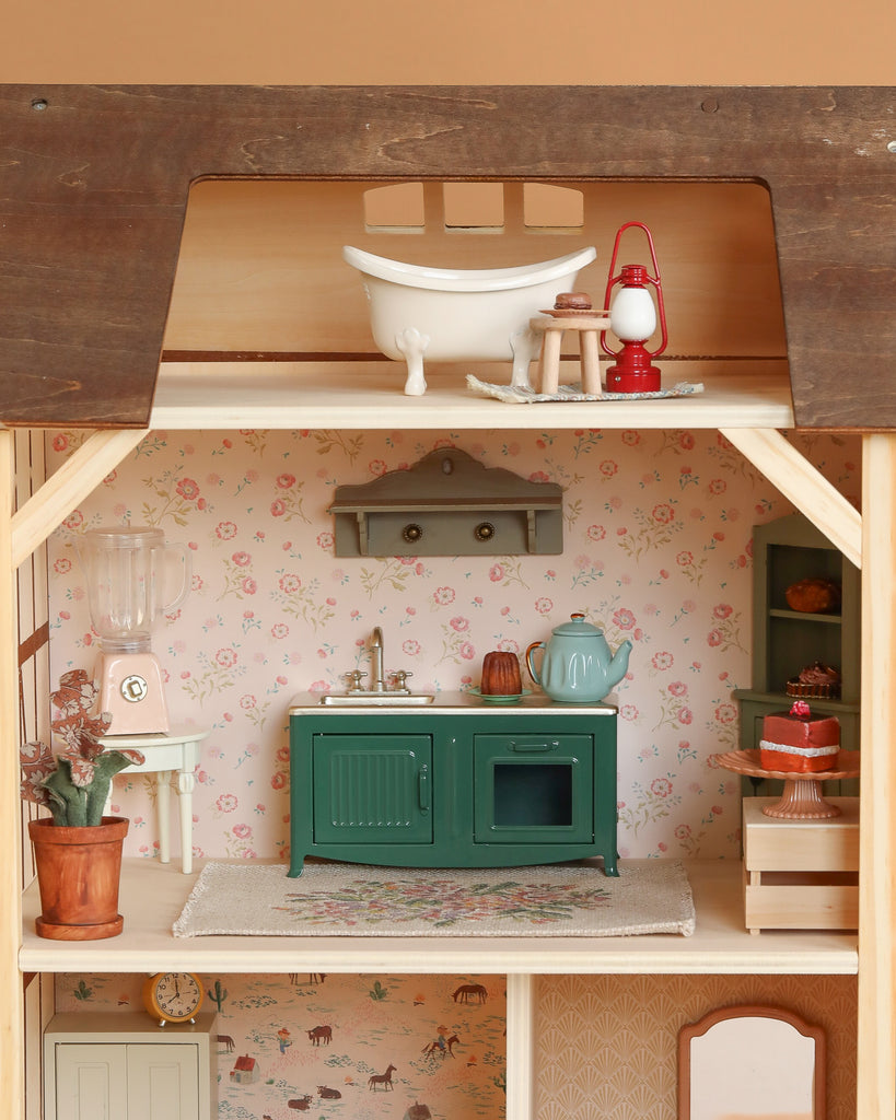 The Maileg Fully Furnished Farmhouse features an upstairs bathroom with a white bathtub and red lantern, plus a kitchen downstairs with floral wallpaper, green cabinet, blue teapot, blender, and charming mini accessories.