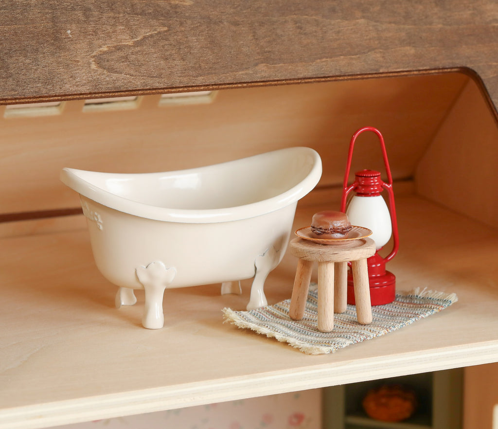 A miniature scene with a white clawfoot tub, red lantern, and stool with glass cup on a striped rug atop wood—perfect for displaying in the Maileg Fully Furnished Farmhouse dollhouse.