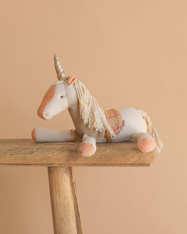 The Maileg Unicorn, Medium—a plush toy with a golden horn, pastel mane and tail, and patterned saddle—rests on a wooden bench against a plain beige background, inviting magical stories.