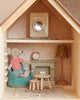 The Maileg Fully Furnished Farmhouse features a cozy room with a stuffed mouse in a striped shirt on a blue chair, plus a table with pastry, mantel clock, lamp, and toy washing machine on the upper shelf.