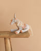 The Maileg Unicorn with Suitcase Set features a soft, light gray unicorn with a pastel mane and tail, sitting on a rustic bench—perfect for collectors or pairing with other Maileg accessories.