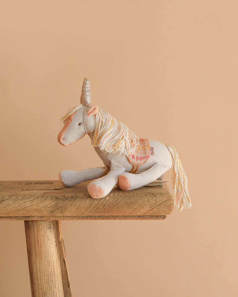 The Maileg Unicorn with Suitcase Set features a soft, light gray unicorn with a pastel mane and tail, sitting on a rustic bench—perfect for collectors or pairing with other Maileg accessories.