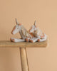 Two Maileg Unicorn, Medium plush toys with beige bodies, orange noses, golden horns, yarn manes, and embroidered saddles sit side by side on a wooden bench against a plain beige background.