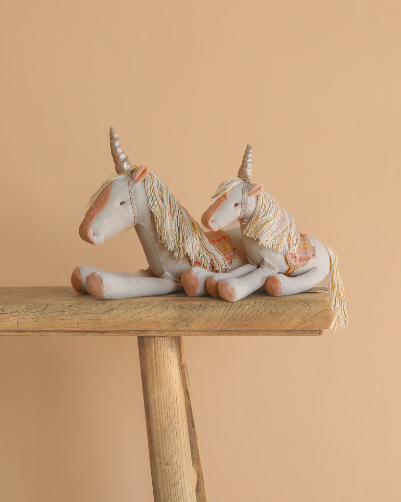 Two Maileg Unicorn, Medium plush toys with beige bodies, orange noses, golden horns, yarn manes, and embroidered saddles sit side by side on a wooden bench against a plain beige background.
