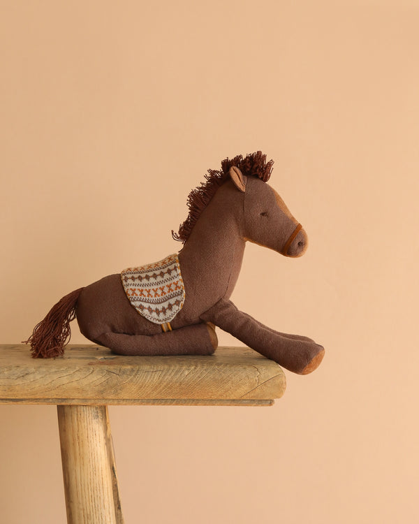 The Maileg Pony, Medium—a brown plush toy made from soft fabric and recycled polyester—rests on a rustic wooden bench set against a plain beige background.