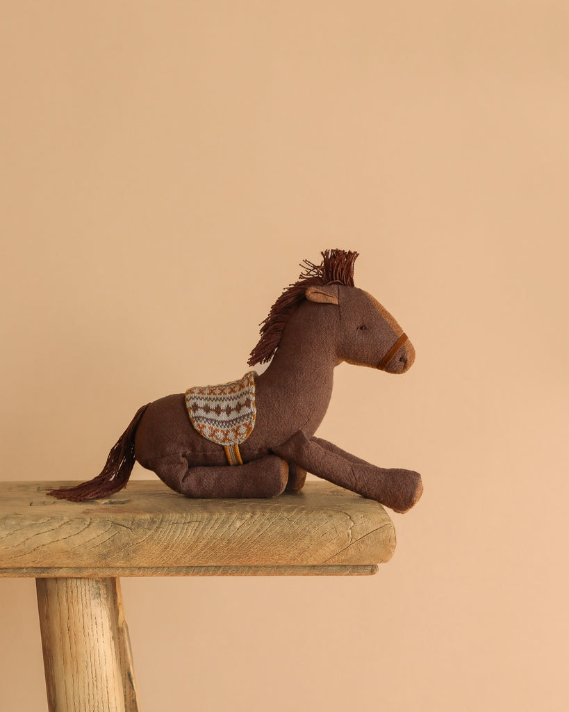The Maileg Cowboy Set, featuring a brown plush horse with a patterned saddle, rests on a wooden bench against a simple beige backdrop.