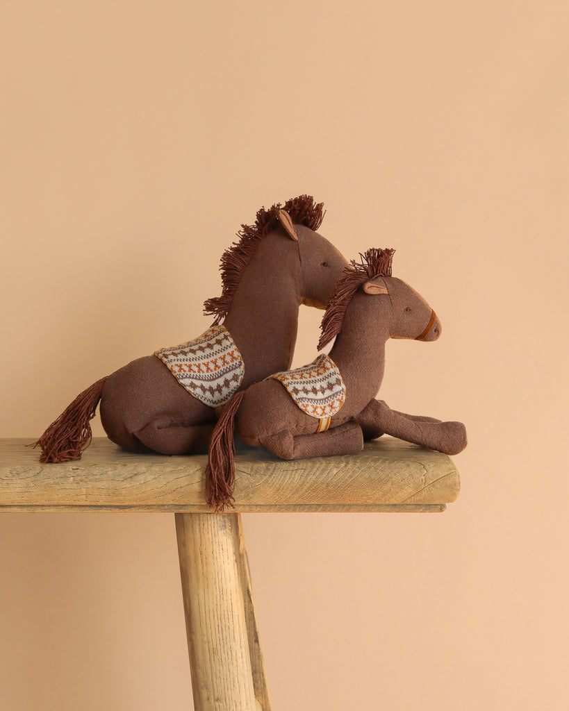 Two Maileg Pony, Medium stuffed toys in brown, made from soft cotton/linen with patterned backs, are displayed on a wooden bench against a plain beige background.