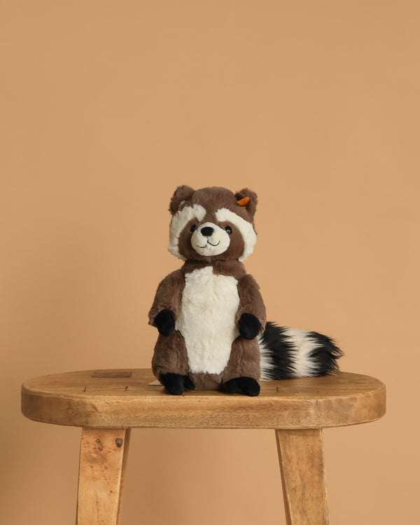 The Steiff Pedro Raccoon 9" is a plush toy with a white belly and striped tail, sitting upright on a wooden stool against a light brown background.