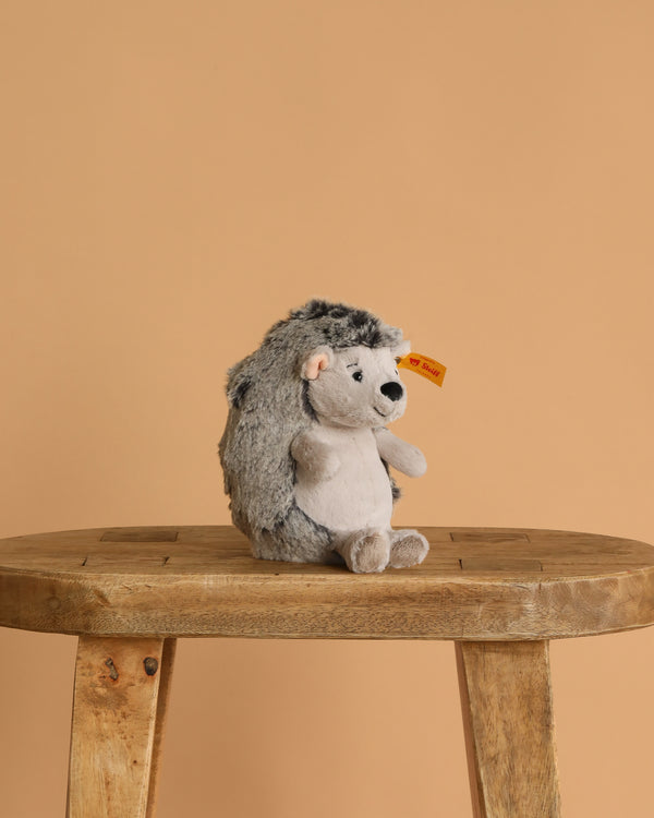 The Steiff Hedgy Hedgehog 6" plush toy with a yellow tag sits upright on a wooden stool against a plain beige background.