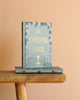 A Christmas Carol | Collector's Edition| Hardcover Book with a blue-green floral cover stands upright on a wooden bench over another copy, set against a beige background, capturing the timeless spirit of Dickens' classic tale.
