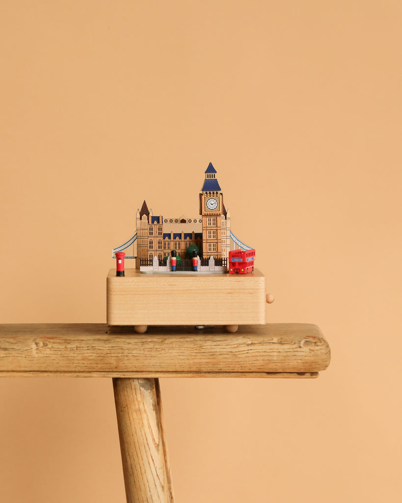 The London Music Box, featuring iconic London landmarks such as Big Ben, a red bus, and a phone booth, sits on a wooden stool against a plain beige background.