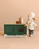 The Maileg Miniature Chef Set features a chef mouse by a green kitchen set, cooking at the stove with a bread loaf and striped rug nearby, set against a beige background.