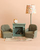 The Maileg Living Room Starter Set includes two green plaid chairs, a teal fireplace with a tiny clock, floral lampshade, and rug—perfectly sized for Maileg mice—all set against a light beige background.