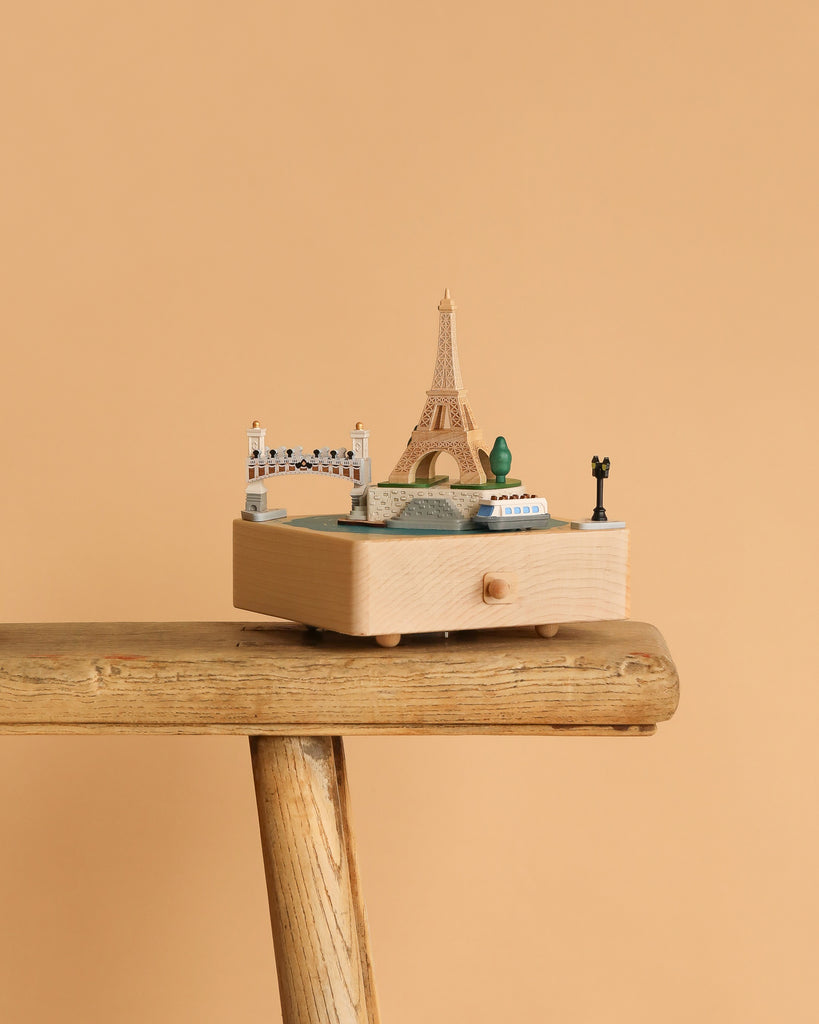 The Paris Music Box, adorned with miniatures of the Eiffel Tower, a bridge, trees, and a boat, is displayed on a rustic wooden stool set against a plain beige background.