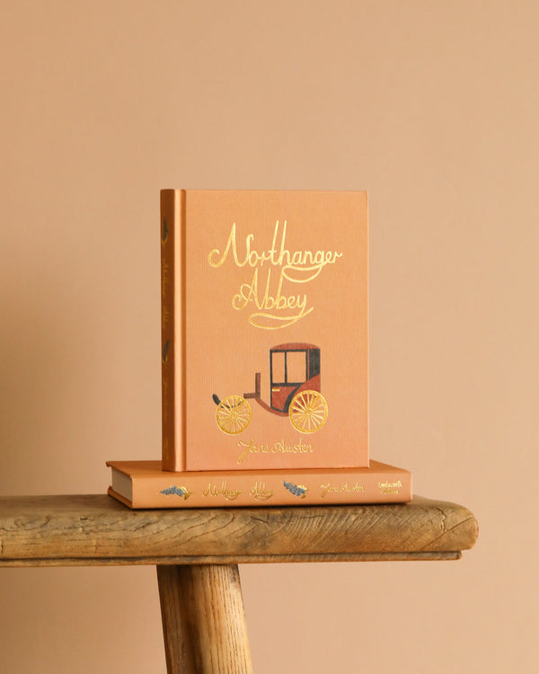 A peach Collector's Edition hardcover of "Northanger Abbey" sits upright on a rustic wooden surface, adorned with gold lettering and a carriage illustration, resting atop another book.