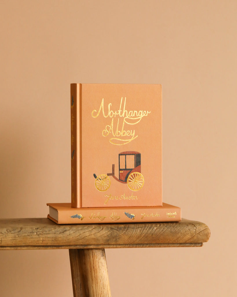A peach Collector's Edition hardcover of "Northanger Abbey" sits upright on a rustic wooden surface, adorned with gold lettering and a carriage illustration, resting atop another book.