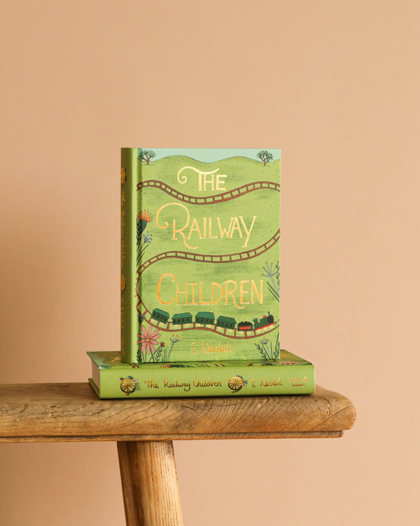 A green hardcover Collector's Edition of "The Railway Children" stands upright on a matching book on a wooden stool, with charming train and plant illustrations on its cover, set against a plain beige background.