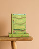 A green hardcover Collector's Edition of "The Railway Children" stands upright on a matching book on a wooden stool, with charming train and plant illustrations on its cover, set against a plain beige background.
