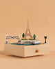 The Paris Music Box displays miniatures of the Eiffel Tower, a riverboat, a bridge, trees, and a street lamp on a plain peach background.