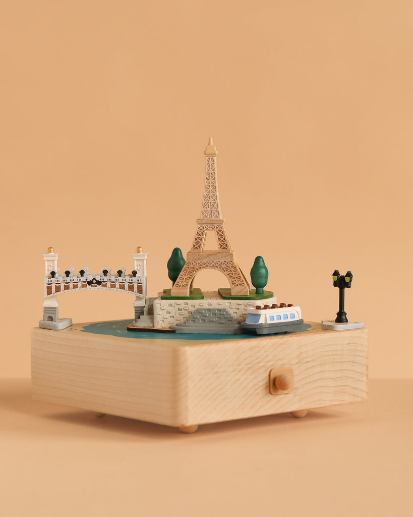 The Paris Music Box displays miniatures of the Eiffel Tower, a riverboat, a bridge, trees, and a street lamp on a plain peach background.