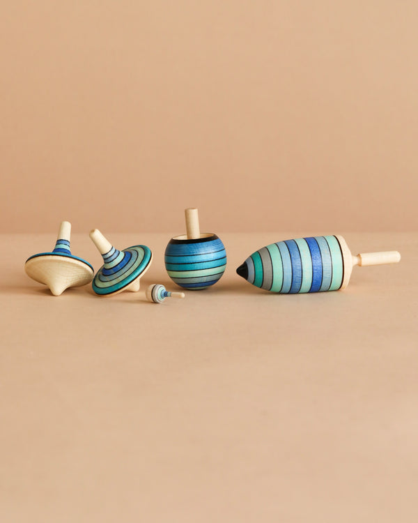 The Mader Spinning Top Champion Learning Set - Ice features five fun tops in various shapes and sizes with blue and green stripes, arranged upright and on their sides on a beige surface—ideal for beginner to advanced spinners.