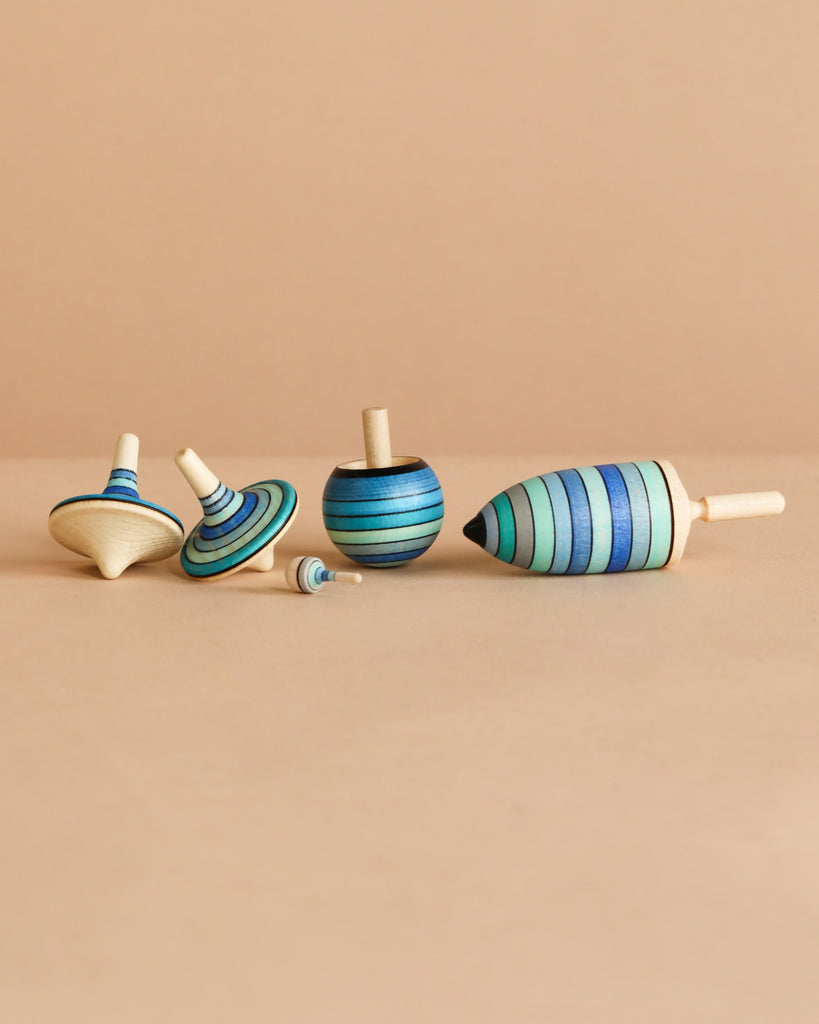 The Mader Spinning Top Champion Learning Set - Ice features five fun tops in various shapes and sizes with blue and green stripes, arranged upright and on their sides on a beige surface—ideal for beginner to advanced spinners.