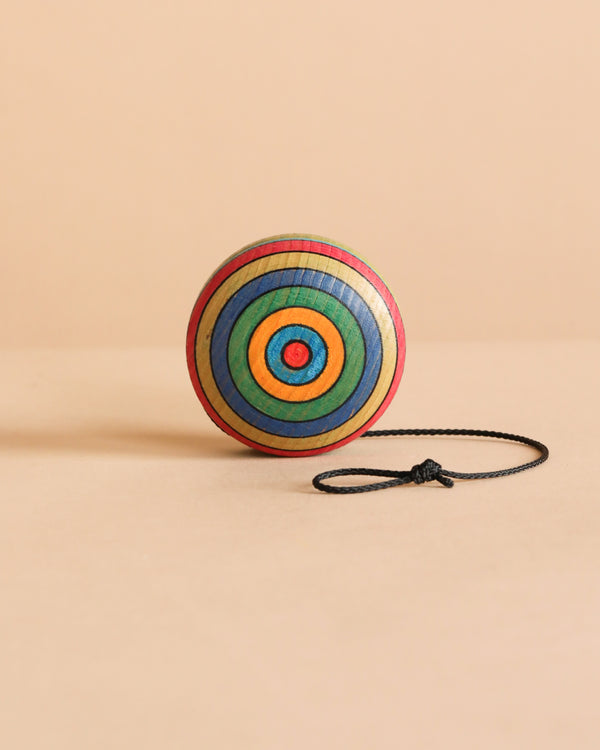 The Mader Wooden Yoyo - Striped, crafted from durable beech wood with colorful concentric circles, stands upright on a beige surface with its black string neatly coiled beside it. This toy meets European safety standard EN71.