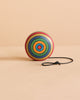 The Mader Wooden Yoyo - Striped, crafted from durable beech wood with colorful concentric circles, stands upright on a beige surface with its black string neatly coiled beside it. This toy meets European safety standard EN71.