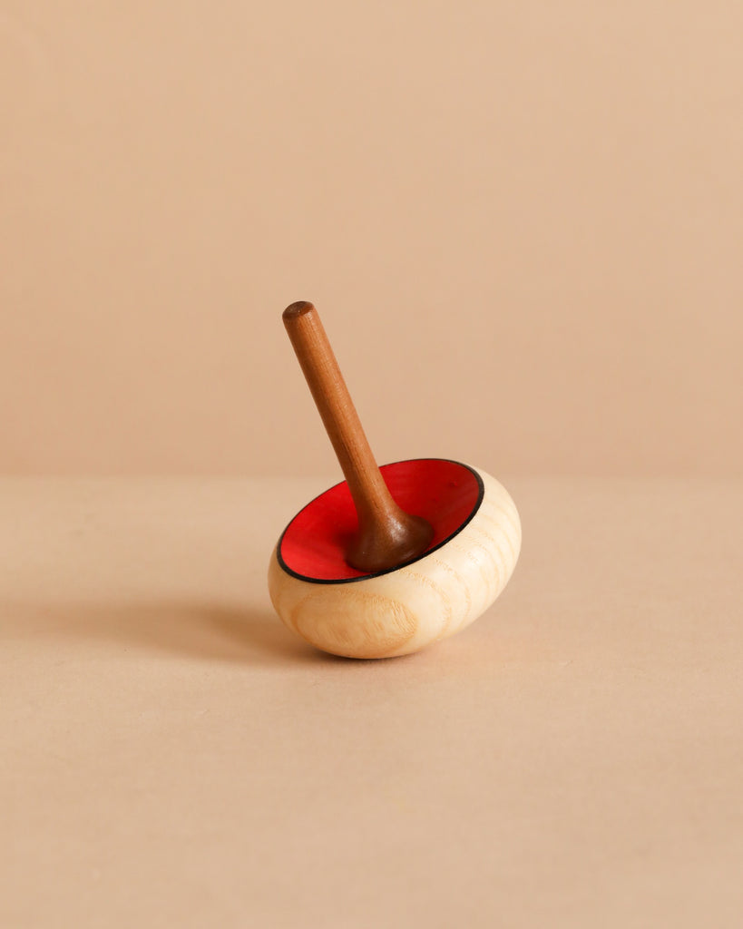 The Mader Zwirbel Spinning Top - Red, crafted from Walnut and Ash with a red interior and natural wood exterior, rests on a beige surface against a matching beige background.