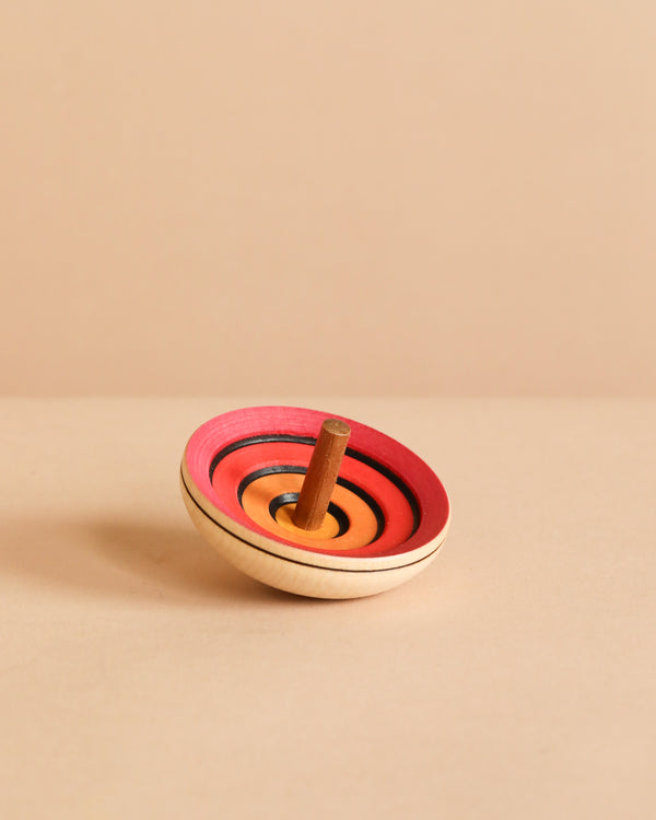 The Mader UFO Flora Spinning Top - Red, made of maple wood with concentric red, orange, and black ombre rings, stands upright on a beige surface with a plain beige background.
