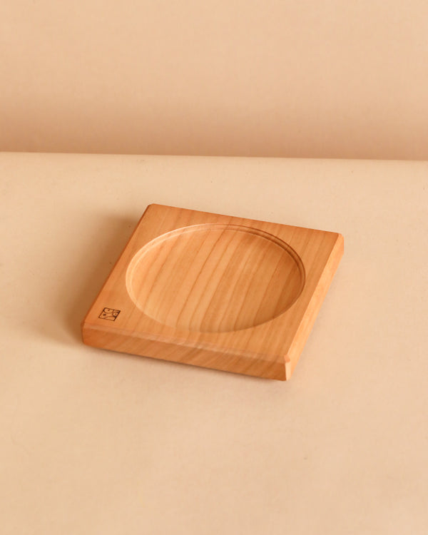 The Cherry Wood Spinning Plate is a square wooden coaster with a circular indented center, featuring a small engraved stamp in one corner. It rests on a beige surface against a soft beige background.