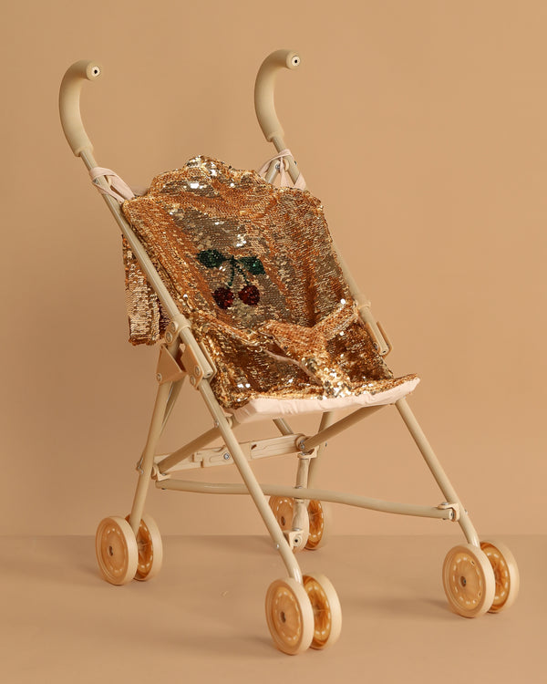 The Konges Sloejd Sparkly Sequin Doll Stroller - Golden Cherry features gold wheels and shiny gold sequin fabric with a cherry design on a matching beige background.