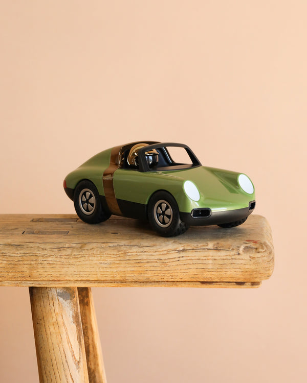 The Playforever Car LUFT HOPPER Green, a small classic racing car toy with a gold stripe, is displayed on a rustic wooden bench with a plain beige background.