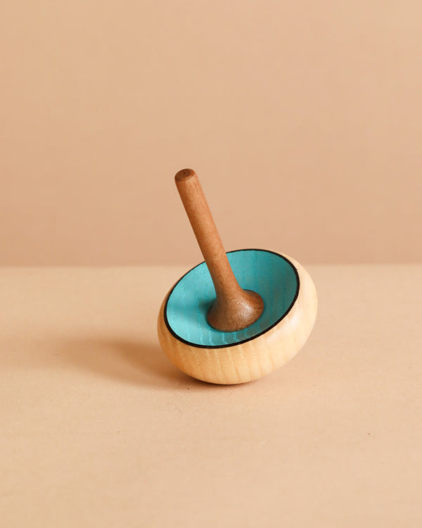 The Mader Zwirbel Spinning Top - Blue, made from Walnut and Ash with a blue interior and natural wood exterior, stands upright on a light beige surface against a beige background.