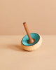 The Mader Zwirbel Spinning Top - Blue, made from Walnut and Ash with a blue interior and natural wood exterior, stands upright on a light beige surface against a beige background.
