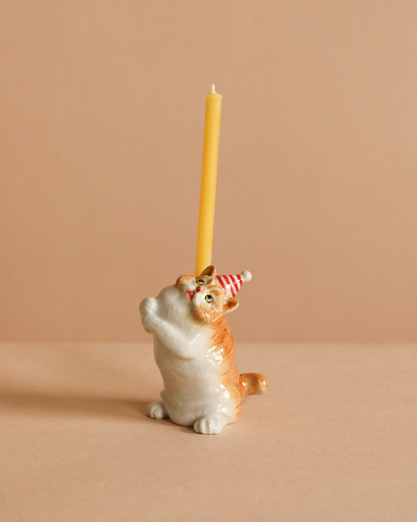 The Exotic Shorthair Cat Cake Topper features a ceramic cat figurine in a red and white party hat, holding a yellow candle upright against a beige background—perfect for adding whimsy to any cat-themed celebration.