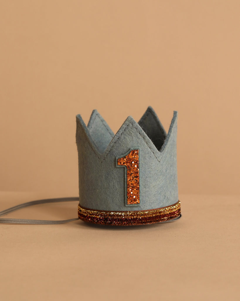 The Storm Gray Felt Crown features a glittery orange number "1" and gold trim at the base—perfect for birthday celebrations—shown against a neutral beige background.