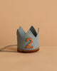 The Storm Gray Felt Crown features a glittery orange number 2 on the front and an orange glitter band around the base—perfect for birthday celebrations—shown against a beige background.