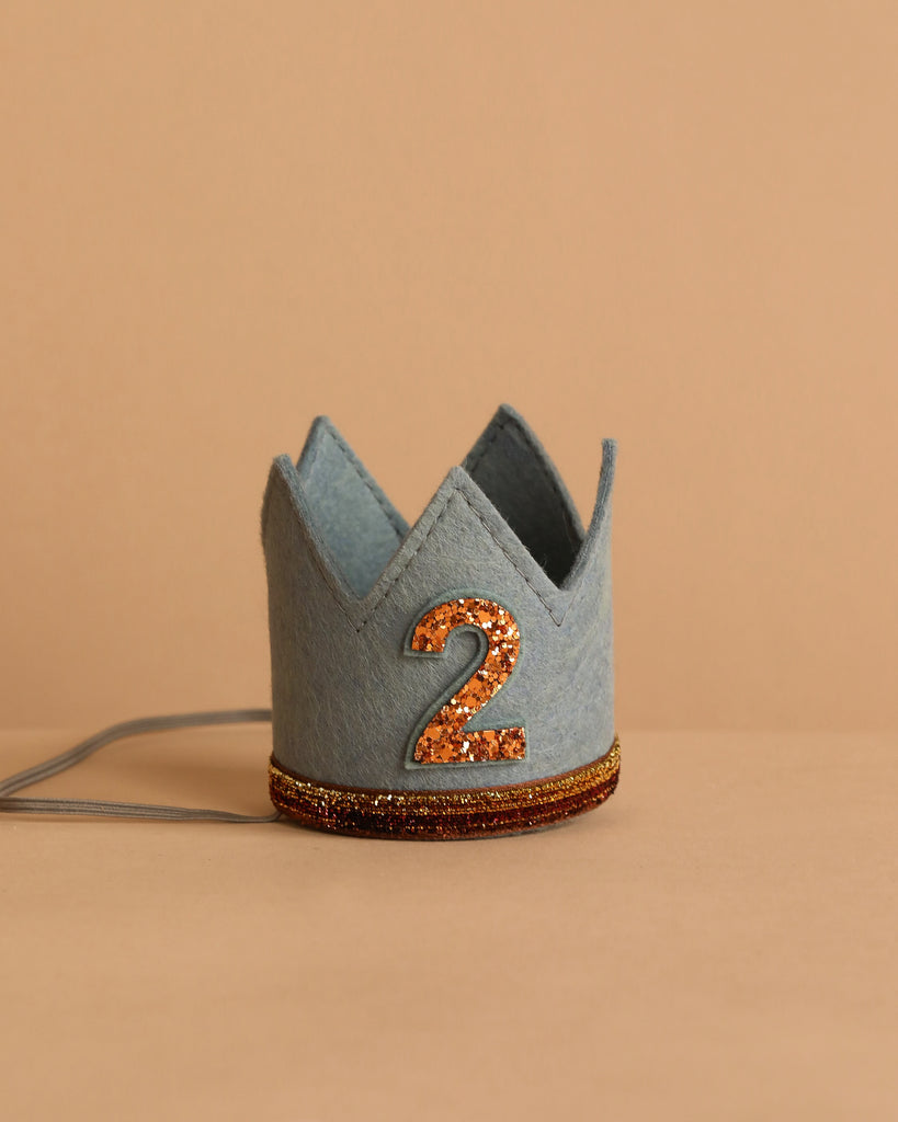 The Storm Gray Felt Crown features a glittery orange number 2 on the front and an orange glitter band around the base—perfect for birthday celebrations—shown against a beige background.