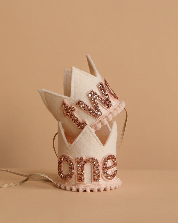 Two small Linen and Rose Crowns, each in cream felt with gold glittery "one" and "two" letters. Perfect party hats for birthdays or photo shoots; feature decorative edges, ribbon ties, and are displayed on a beige surface.