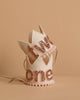 Two small Linen and Rose Crowns, each in cream felt with gold glittery "one" and "two" letters. Perfect party hats for birthdays or photo shoots; feature decorative edges, ribbon ties, and are displayed on a beige surface.